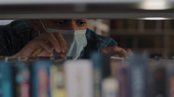 Closeup Hispanic Guy Student in Protective Medical Mask Looking for Right Book on Bookshelf in alt