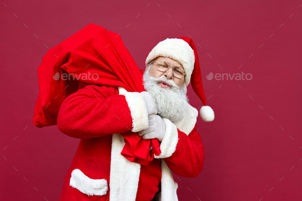 Tired Santa Claus feeling back pain holding heavy bag on red background.