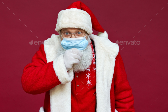 Sick ill Santa Claus wearing face mask coughing isolated on red background.
