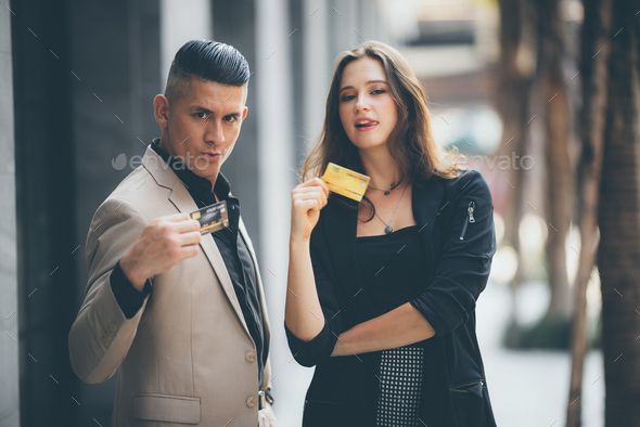 Young stylish male and female holding credit card with facial expression standing at footpath