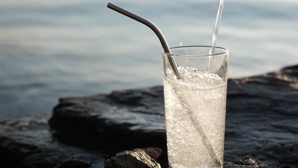 Soda Water Pours Into Tall Glass with Reusable Cocktail Tube on Rocky Seashore alt