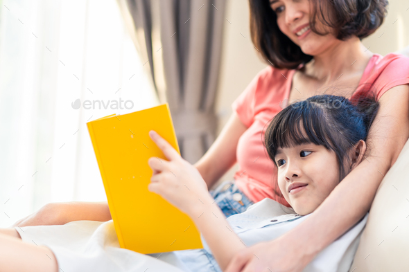 Asian beautiful mother and her daughter reading fairy tail story book on sofa at home together
