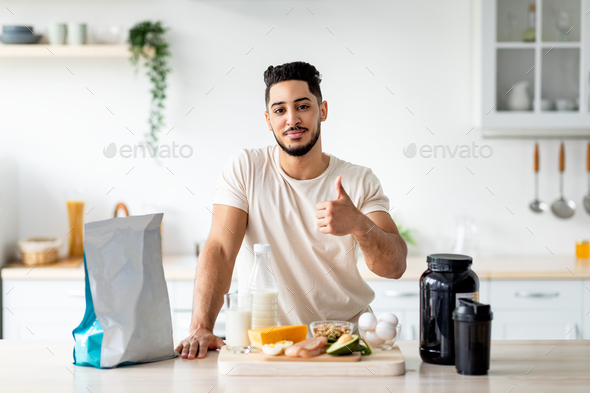 Sporty young Arab guy with wholesome foods and protein shake gesturing thumb up at kitchen - Stock Photo - Images