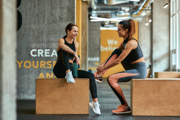 Full length of two young athletic fitness girls in sportswear sitting on crossfit jump boxes at gym