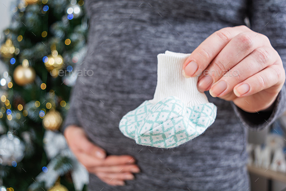 Young pregnant woman with swollen belly and baby socks in her hand posing against Christmas tree