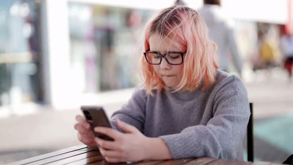 A Teenage Girl with Glasses is Sitting at a Table in a Street Cafe and Playing Video Games on Her alt