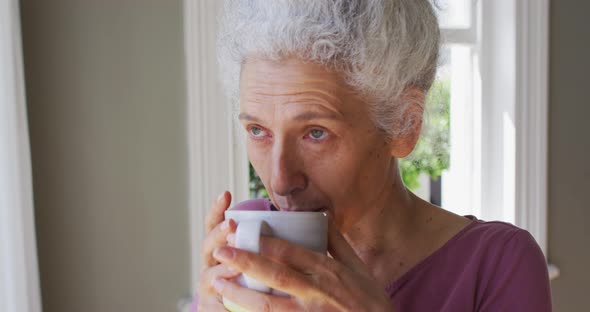 Close up of caucasian senior woman drinking coffee while looking out of the window at home alt