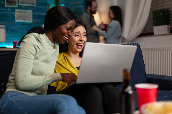 Multi-ethnic women sitting on sofa holding laptop computer looking at comedy entertainment video