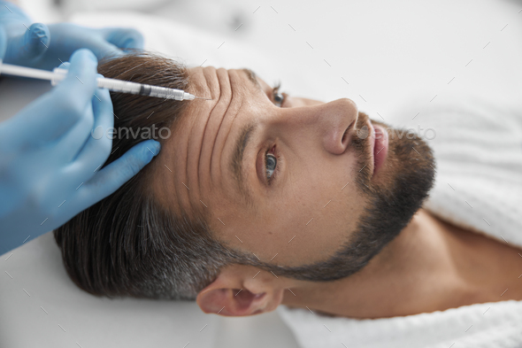 Doctor cosmetologist does injection of lifting filler in wrinkled forehead of mature man in clinic