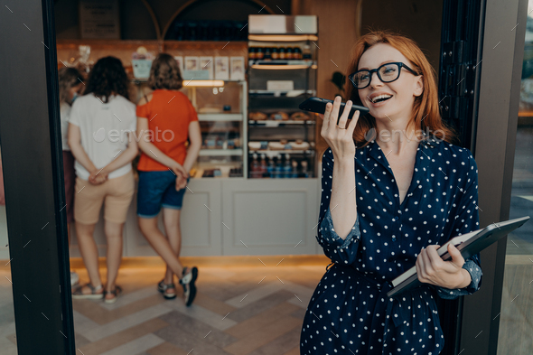 Redhead young woman makes voice call keeps smartphone near mouth wears spectacles polka dot dress
