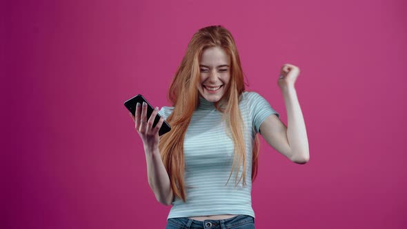 The Young Winner with the Phone in Her Hand Says Wow and Smiles Happily in a Blue Casual Tshirt alt
