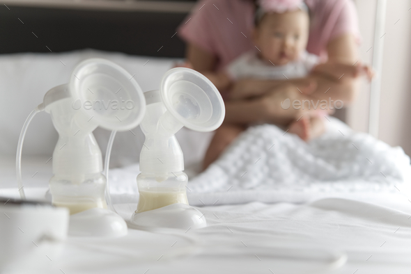 Breast milk pump\'s bottles and pump machine on the bed with mother hugging baby in background.