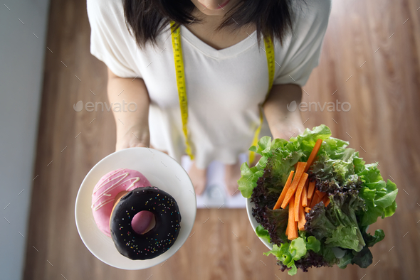 Top view of woman holding dishes of vegetable salad and donuts standing on weight measurement scale