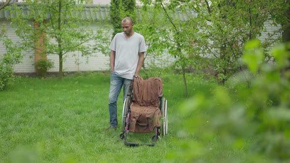 Wide Shot of Thoughtful Adult African American Caregiver Standing at Wheelchair on Summer Backyard alt