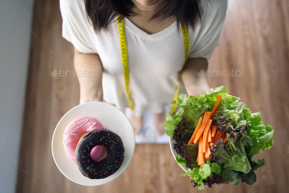 Asian girl holding vegetable salad and donuts standing on weight measurement scale
