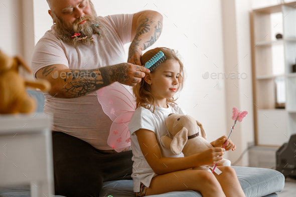 Daddy with scrunchies in beard brushes hair of cute little daughter at home
