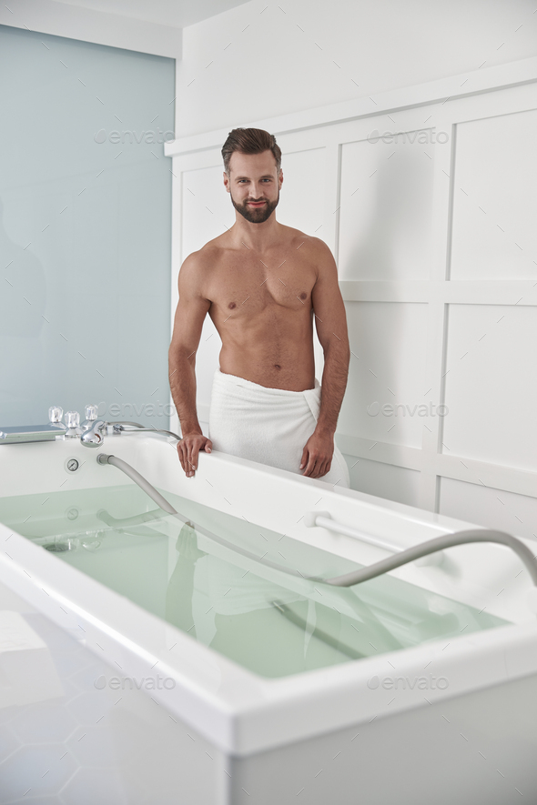 Athletic man with bare chest stands by modern hydro massage tub in clinic