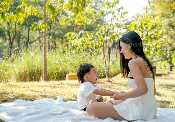 Young Asian mother play and fun with her little boy on white carpet in the green garden