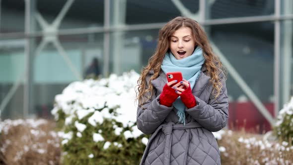 Young Beautiful Curly Woman in Winter Clothes Holding Phone Celebrate Good Mobile News Surprise Bid alt