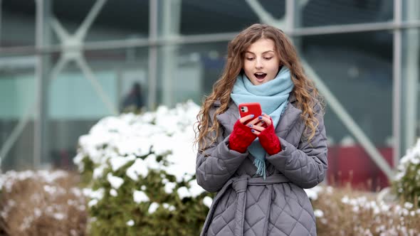 Young Beautiful Curly Woman in Winter Clothes Holding Phone Celebrate Good Mobile News Surprise Bid