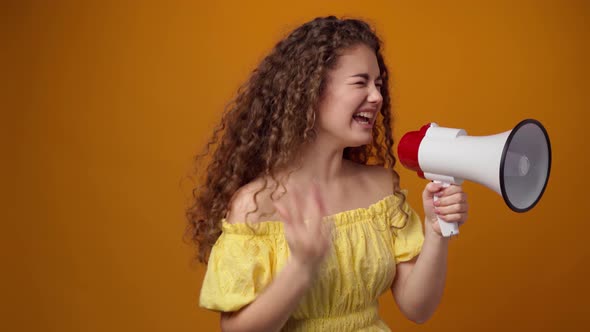 Curlyhaired Young Woman Shouting Into Loudspeaker Against Yellow Background