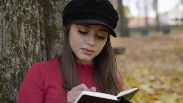 Thoughtful Young Girl Writing Down in a Notebook in in the Autumn Park alt