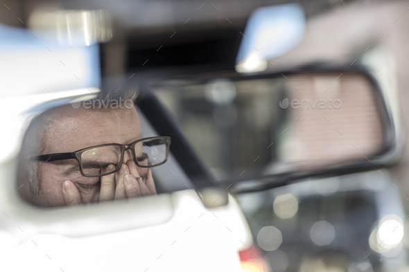 Reflection on man in rear view mirror in car, rubbing tired eyes