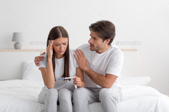 Sad unhappy young caucasian husband calm his wife on bed, lady look at pregnancy test in bedroom