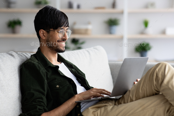Positive indian guy chilling at home, using laptop