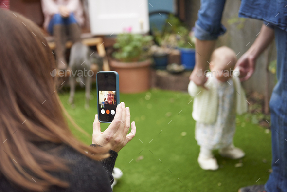 Mother in garden using smartphone to film daughter learning to walk, focus on foreground