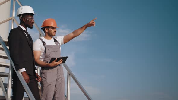 Indian Engineer in Orange Helmet and Grey Overalls Pointing in Distance and alt