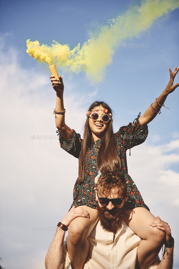 Young boho woman holding yellow smoke flare on boyfriend\'s shoulders at festival