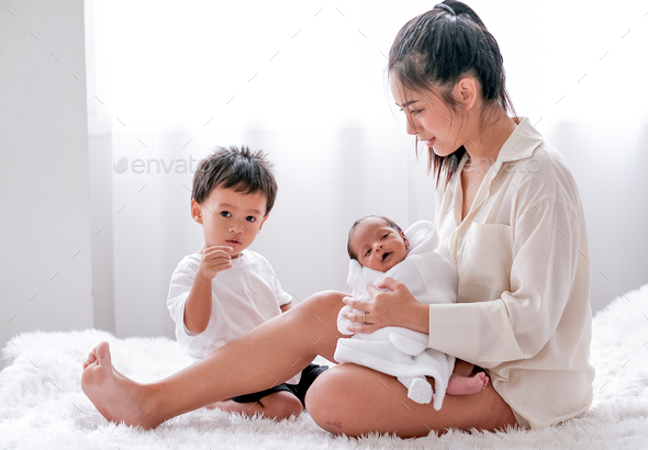 Asian mother carry her newborn baby on lap and sit on bed with her son in the bedroom. - Stock Photo - Images