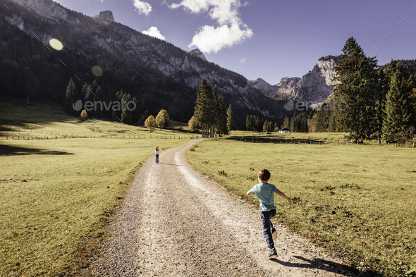 Rear view of boy running on dirt track toward girl in valley landscape, Bavaria, Germany