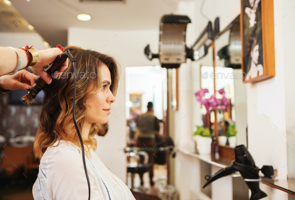 Hairdresser using curling tongs on customer\'s long brown hair in salon