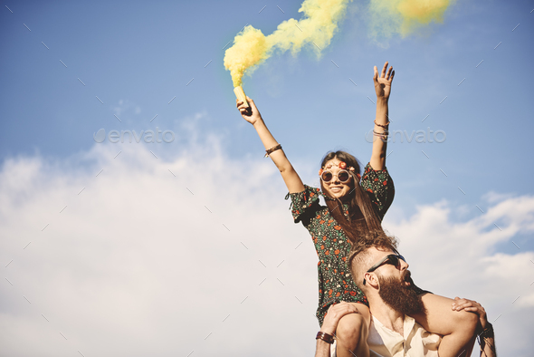 Young boho woman holding yellow smoke flare on boyfriend\'s shoulders at festival