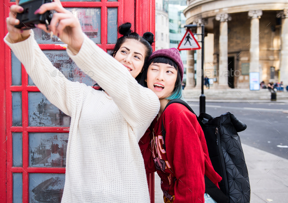 Two young stylish women taking selfie by red phone box, London, UK - Stock Photo - Images