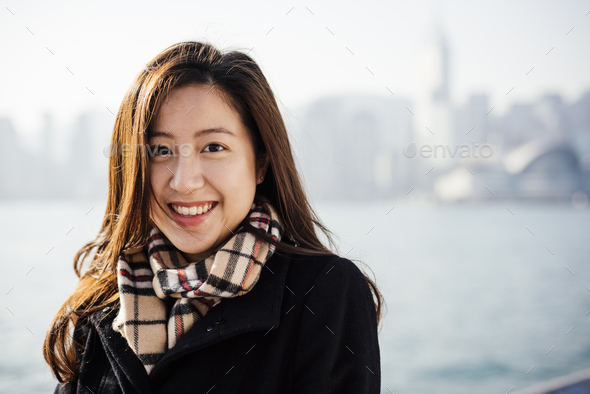 Portrait of young woman wearing checked scarf in front of water, looking at camera smiling