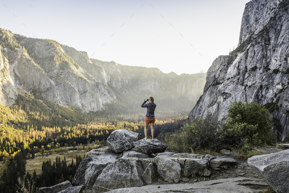 Woman on boulder looking out at valley forest through binoculars, Yosemite National Park, California