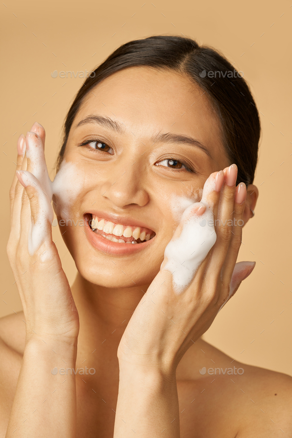 Beauty portrait of joyful young woman smiling at camera while applying gentle foam facial cleanser