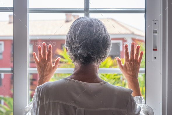 Rear view of mature woman gray haired looking out of the window with hands on glass