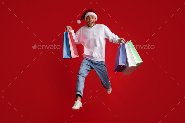 Christmas Sales. Funny Excited Man Wearing Santa Hat Jumping With Shopping Bags