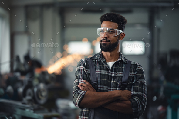 Portrait of young industrial man standing with arms crossed indoors in metal workshop, looking away