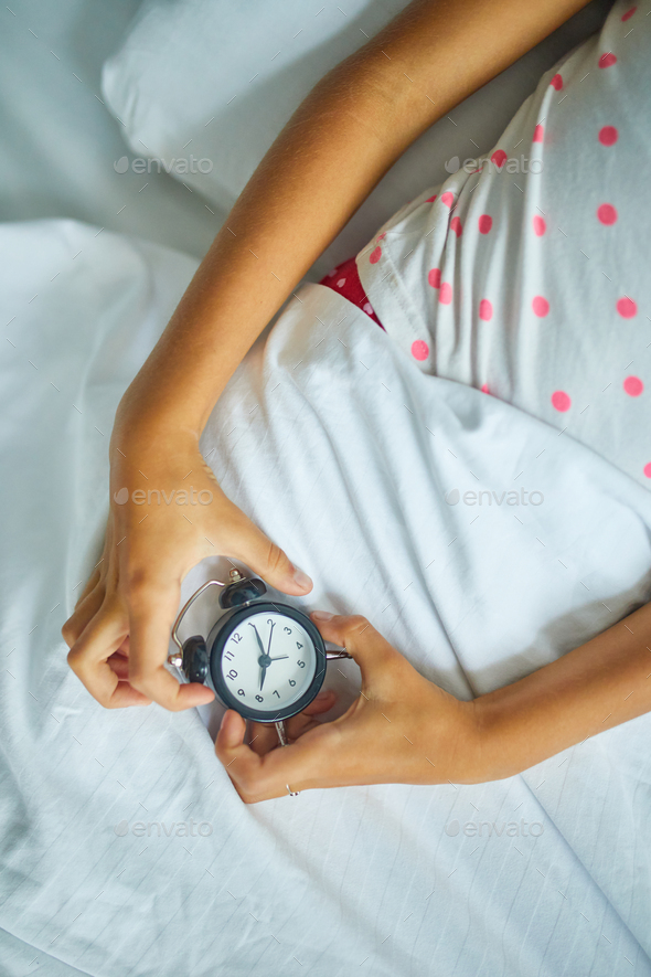 Little girl hold in hand an alarm clock, wakes up from sleep on a big and cozy bed