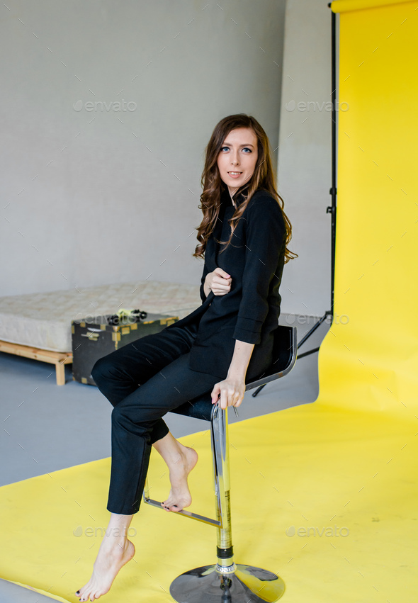 girl with long dark hair in a black suit sits on a bar stool, yellow background, in the studio.