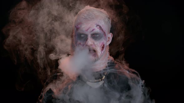 Frightening Man with Halloween Zombie Makeup Blows Smoke From Nose and Mouth Trying to Scare alt