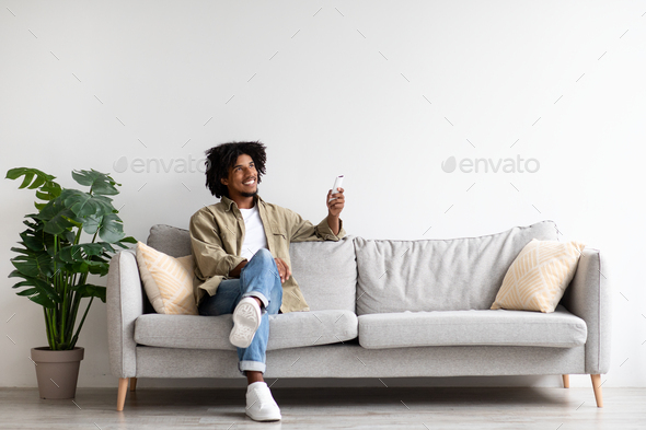 Happy Black Guy Sitting On Couch, Operating Air Conditioner With Remote Controller
