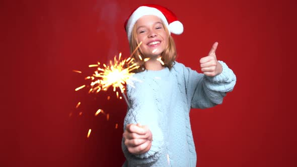 Happy Little Girl in Santa Claus Cap Holding Bengal Light on the Red Background