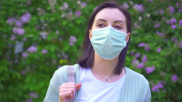 Portrait of a Young Woman in a Medical Mask on the Background of Flowering Plants in the Park alt