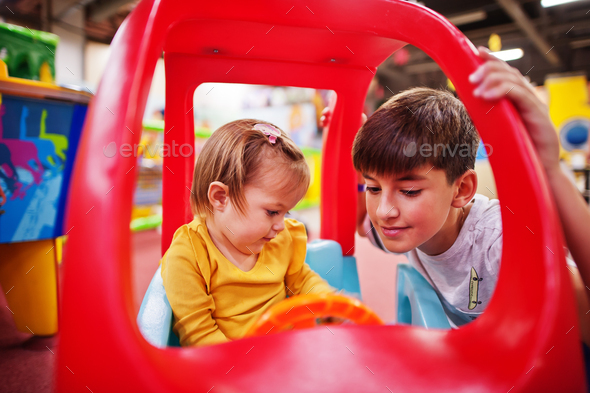 Brother with sister rides on a plastic car in indoor play center.
